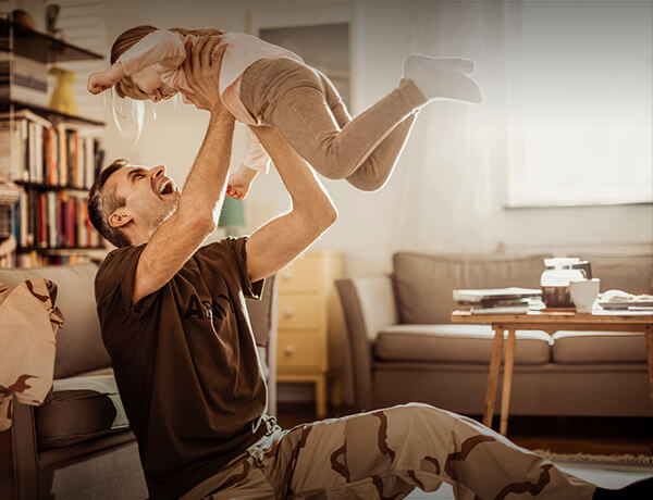 Man lifting a child into the air while sitting on the floor in camo fatigue pants