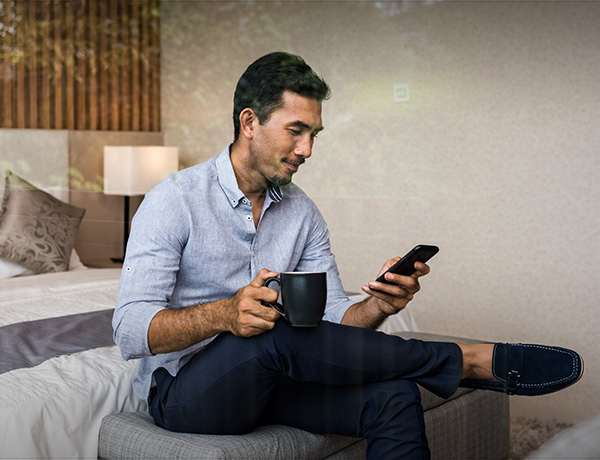 Man sits on hotel bed holding coffee mug and looking at cell phone